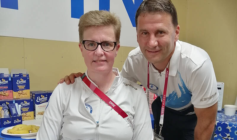 Mies kumartuneena naisen vierelle, pitäen kättä tämän olkapäällä. Molemmat hymyilevät kameralle.