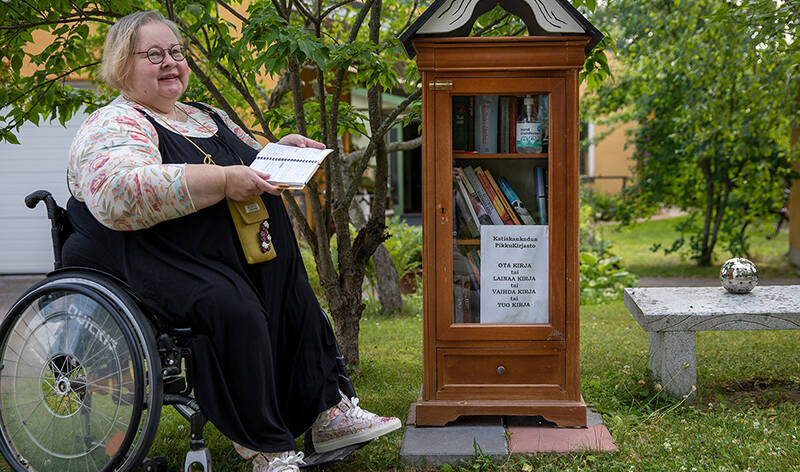 Nainen istuu pyörätuolissa pensaan vierellä ja pitää kädessään avoinna olevaa kirjaa. Vieressä on pieni lasiovinen kaappi, jossa kirjoja sekä kaapin vieressä on kivipenkki.