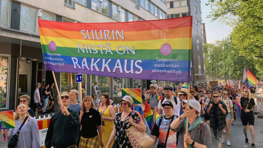 Ihmiset kävelevät kulkueessa ja he kantavat kylttiä, joka on sateenkaaren värinen ja siinä lukee Suurin niistä on rakkaus.