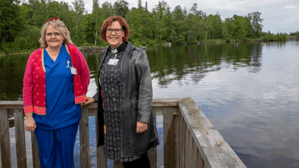 Kaksi naista nojaavat laiturin kaiteeseen ja hymyilevät. Tyyni järvi ja vihreä metsä näkyvät heidän takana.