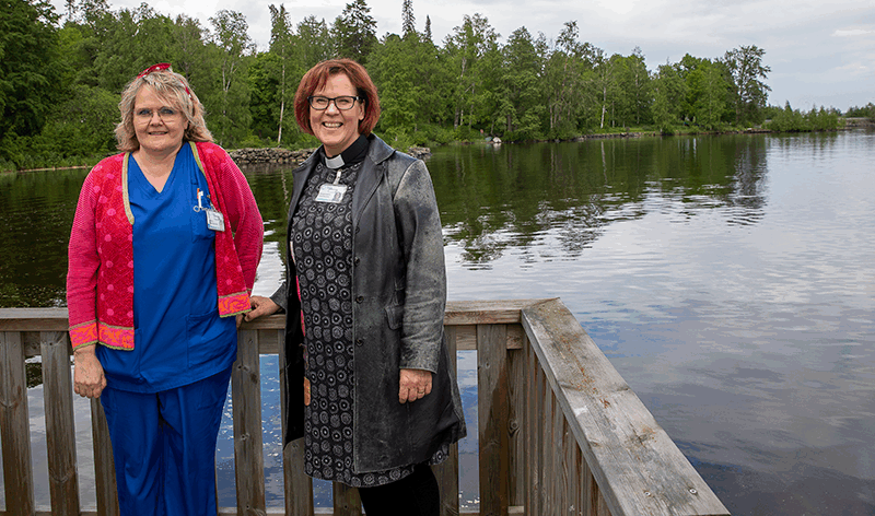 Kaksi naista nojaavat laiturin kaiteeseen ja hymyilevät. Tyyni järvi ja vihreä metsä näkyvät heidän takana.