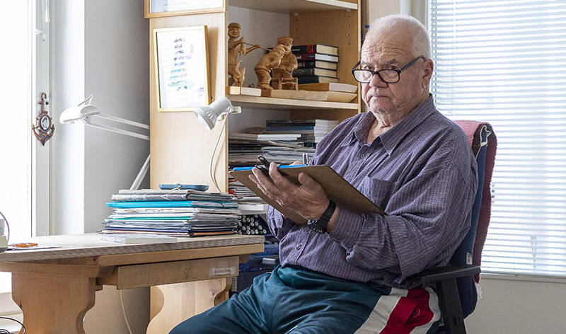 Vanha mies istuu työpöydän ääressä käsissään taustapahvin päällä oleva lehtiö sekä kynä.