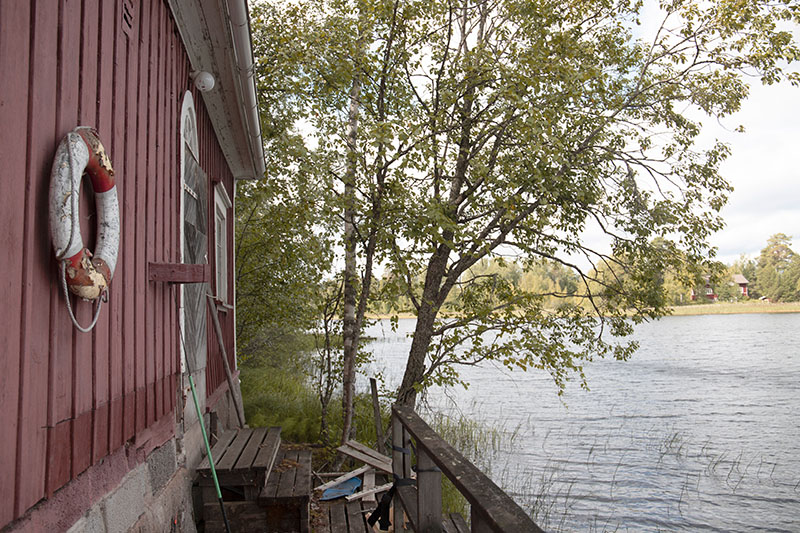 Vasemmalla näkyy punaista rakennusta, jonka seinällä on vanha pelastusrengas. Oikealla on järveä, puu ja laituri.