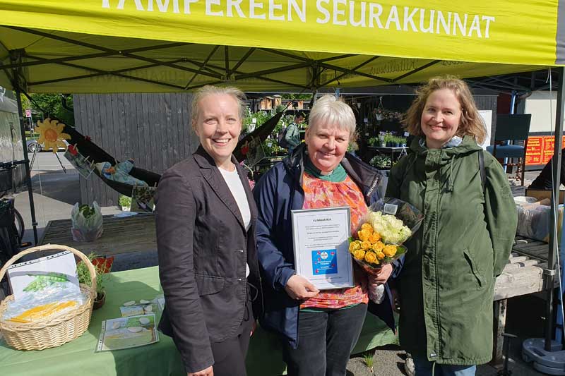 Kolme hymyilevää naista seisoo Tampereen seurakuntien telttakatoksen edessä ja keskellä olevalla on kehystetty kunniakirja käsissään.