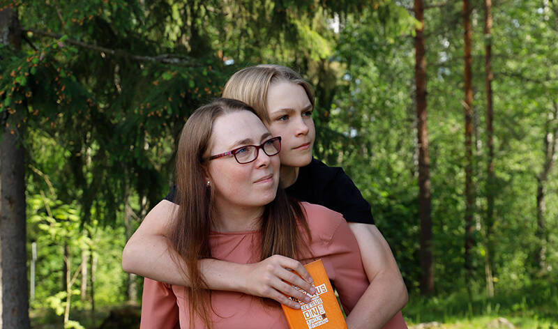 Äiti pitää reppuselässä rippikouluikäistä poikaa metsässä. Molemmat katsovat samaan suuntaan sivulle ja pojalla on kädessään oranssikantinen raamattu.