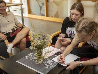 Yksi ihminen istuu rennosti ja kaksi muuta kirjoittaa jotain muistiin pöydän ääressä. Kirjoittajilla on paidoissaan teksti All Inn.