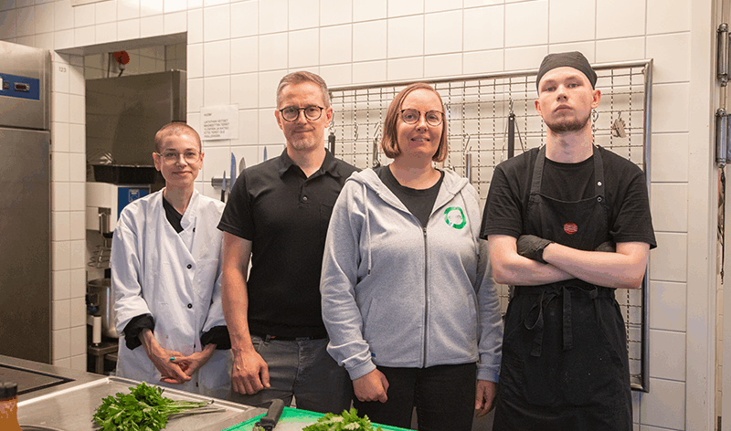 Neljä henkilöä seisoo rivissä laitoskeittiössä ja katsovat kameraan. Kuvan edessä näkyy kaksi leikkuulauttaa joissa on salaattia.
