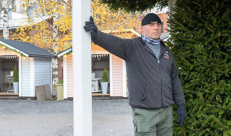 Mies nojaa kädellään tukipylvääseen, taustalla on pieniä mökkejä.