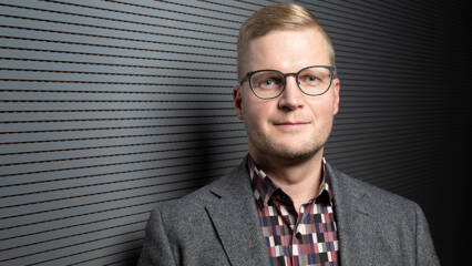 Ylävartalokuva henkilöstä, jolla on vaalea pukutakki ja ruudullinen paita.