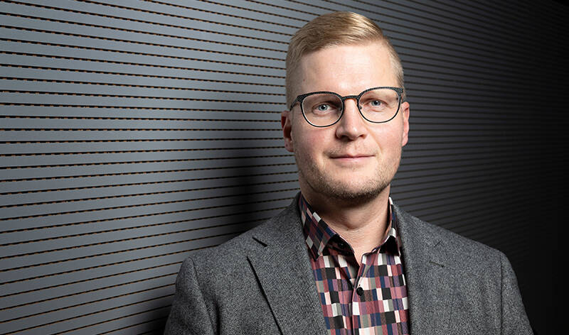 Ylävartalokuva henkilöstä, jolla on vaalea pukutakki ja ruudullinen paita.