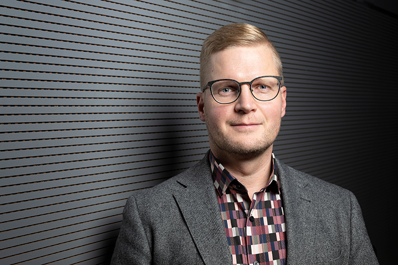 Ylävartalokuva henkilöstä, jolla on vaalea pukutakki ja ruudullinen paita.