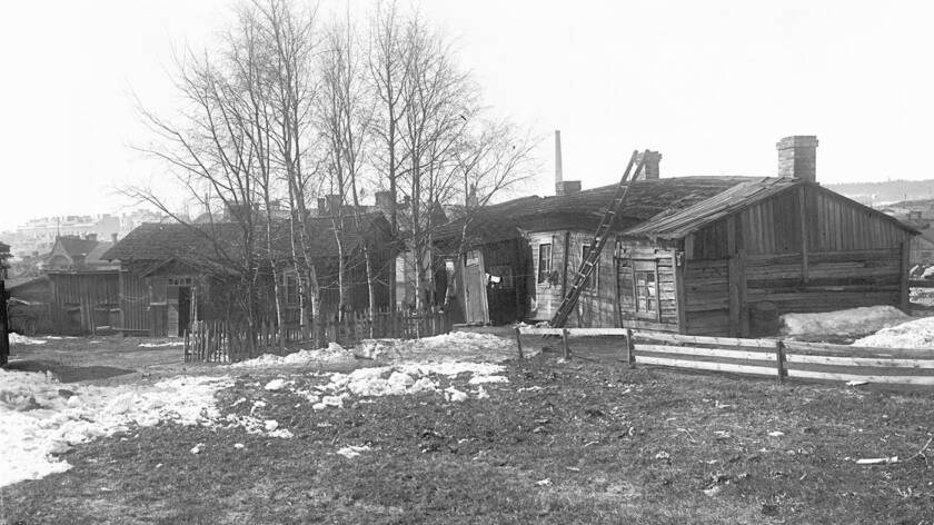 Mustavalkoinen kuva, jossa vanha 1900-luvun puutalo. Pihassa lehdettömiä puita ja sekä aitoja. Taloa vasten nojaa tikapuut.