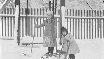 1900-luvun alkupuolen mustavalkoisessa kuvassa kaksi tyttöä talvisissa puuhissa. Toinen hiihtää ja toinen potkukelkkailee.