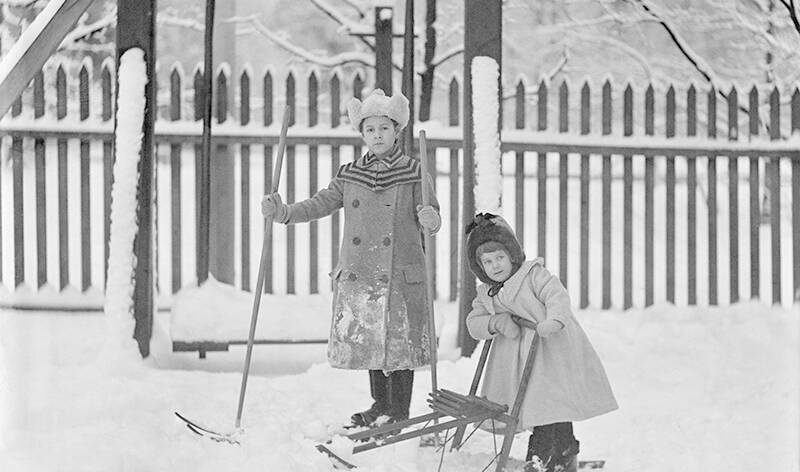 1900-luvun alkupuolen mustavalkoisessa kuvassa kaksi tyttöä talvisissa puuhissa. Toinen hiihtää ja toinen potkukelkkailee.