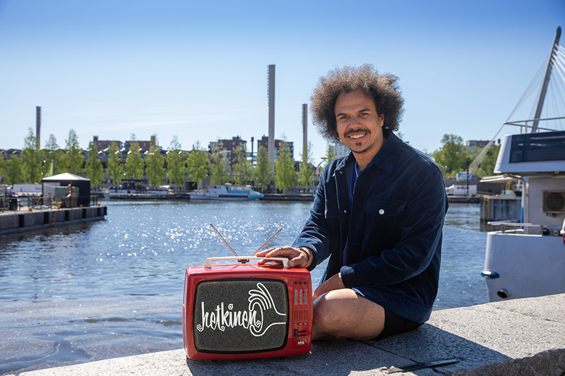 Naurava mies istuu shortsit jalassa veden äärellä. Vieressä reunavallilla on vanha minitelevisio, jossa lukee hetkinen.