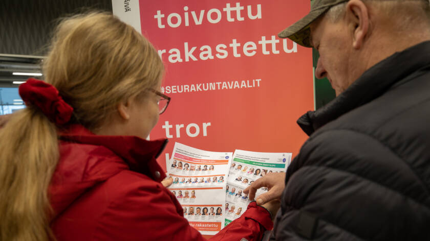 Mies ja nainen pitävät käsissään kuvallisia ehdokaslistoja ja katsovat niitä, taustalla roll-up, jossa lukee uskottu, toivottu, rakastettu.