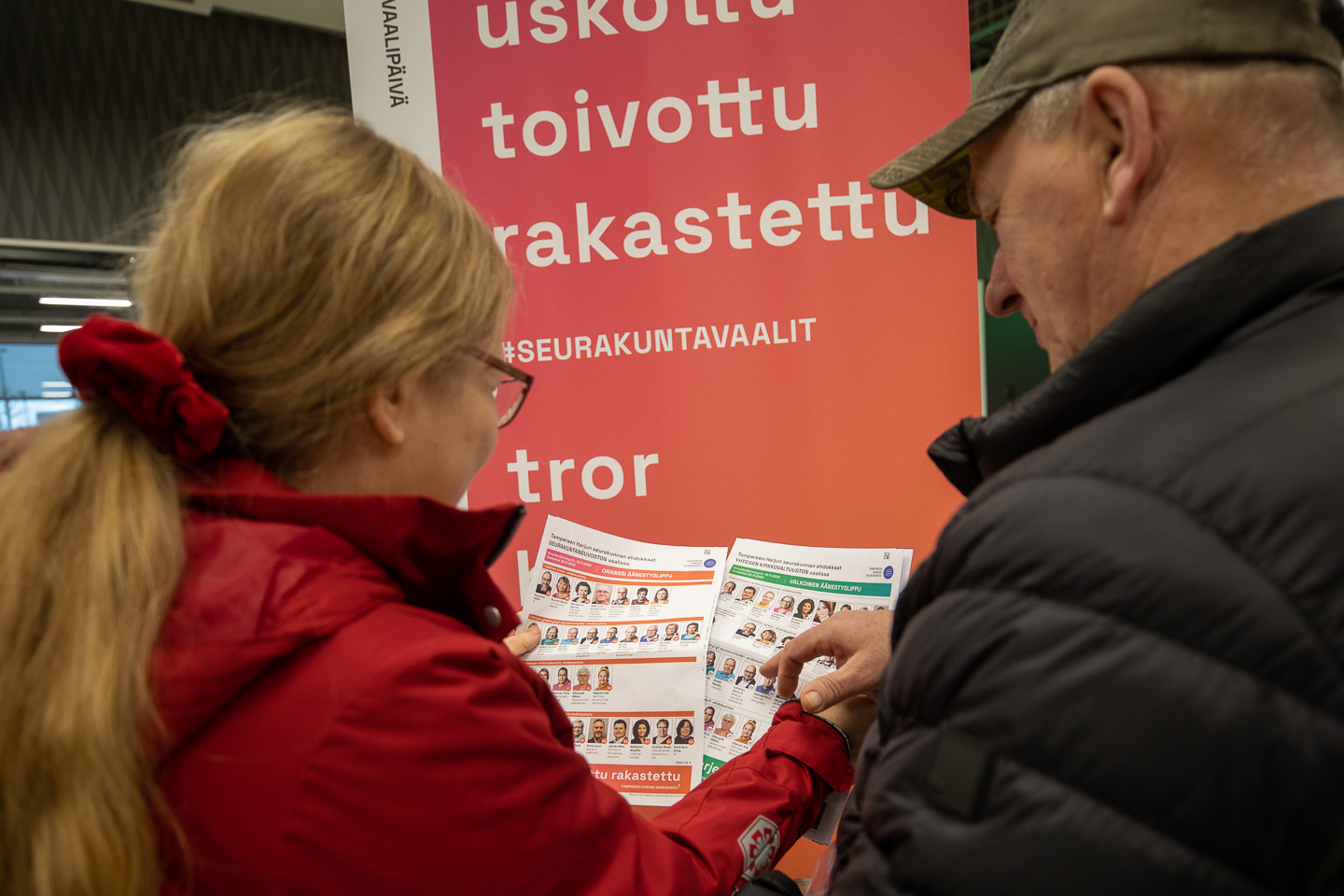 Mies ja nainen pitävät käsissään kuvallisia ehdokaslistoja ja katsovat niitä, taustalla roll-up, jossa lukee uskottu, toivottu, rakastettu.