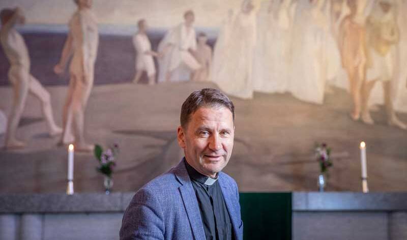 Henkilö, jolla on harmaa puku ja papin panta seisoo Tampereen tuomiokirkon alttaritaulun edesså, jossa on ryhmä ihmisiä, jotka kävelevät kuvan oikealle puolelle.