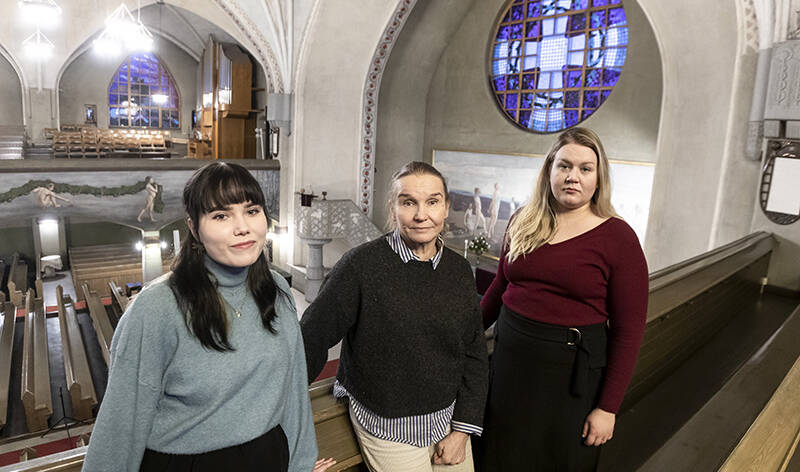 Kirkon parvella seisoo kolme naista, joiden taustalla kirkon sisätilat levittäytyvät laajana näkymänä.