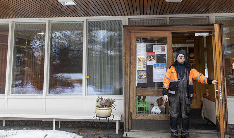 Henkilö seisoo työhaalareissa keskellä rakennuksen ovensuuta pitäen ovea auki. Oven ympärillä on useita ikkunoita.