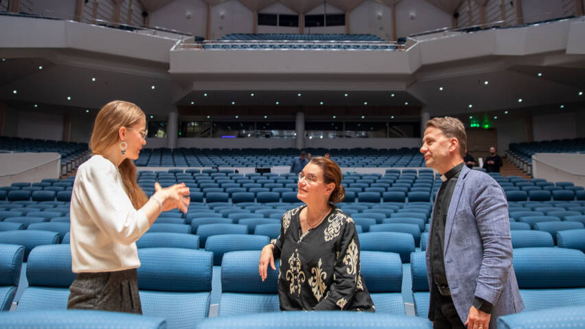 Suvi Leinonen, Hanna Suutela ja Olli-Pekka Silfverhuth seisovat suuren katsoman sinisten penkkien välissä keskustellen.