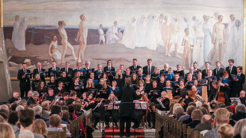Noin kolmenkymmenen hengen sekakuoro seisoo ihmisä täynnä olevan kirkon alttarioen edessa ja heidän edessään soittaa sinfoniaorkesteri.