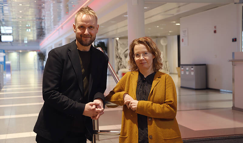 Kaksi ihmistä nojaa keskellään olevaan kaiteeseen sisällä valoisassa hallitilassa.