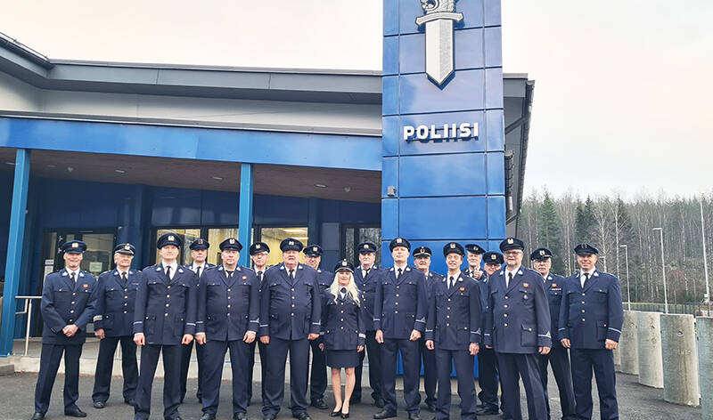 Iso joukko sinisiin univormuihin ja koppalakkeihin pukeutuneita ihmisiä seisoo jämäkästi ulkona rakennuksen edessä. Rakennukseen liittyy pystysuuntainen kyltti, jossa on sana poliisi sekä tunnuskuva miekasta, jonka yläpäässä on leijona kruunu päässään.