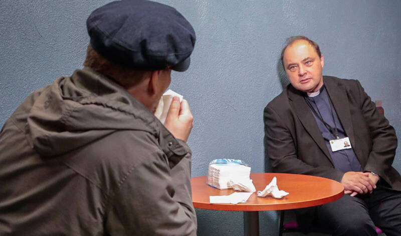 Kaksi miestä, joista toinen on pappi, istuvat pyöreän pöydän ääressä.