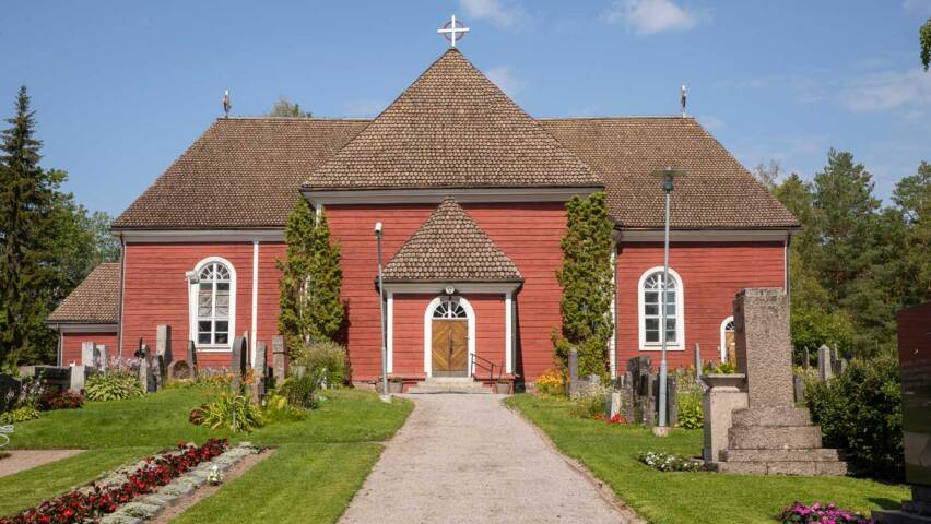 An old red wooden church with a grey shingle roof in a summery landscape.