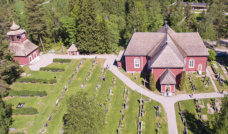 Ilmakuvassa on punainen puukirkko, kellotapuli ja hautausmaata.