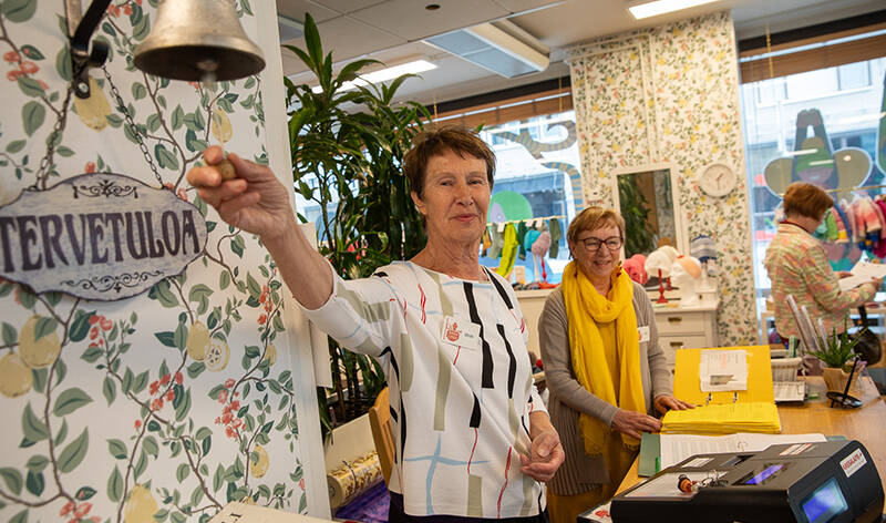 Yksi ihminen kurkottaa kilistäen kelloa, joka on Tervetuloa-kyltin yläpuolella. Taustalla toinen seisoo tiskin takana, jolla on kassakone ja mappi. Taustalla on isot ikkunat, ihminen ja käsitöitä.