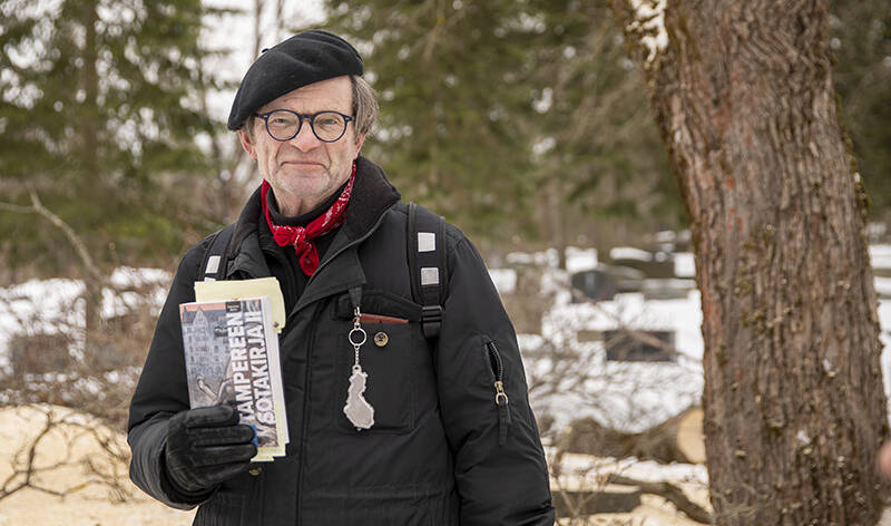 Talvinen kuva miehestä hautausmaalla kirja kädessään. Vieressä on puunrunko.