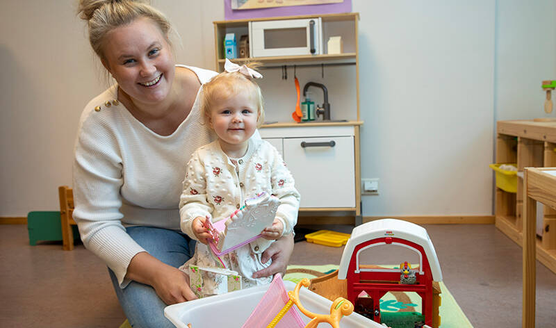 Nainen hymyilee lapsen kanssa kameralle lelukorin takaa.