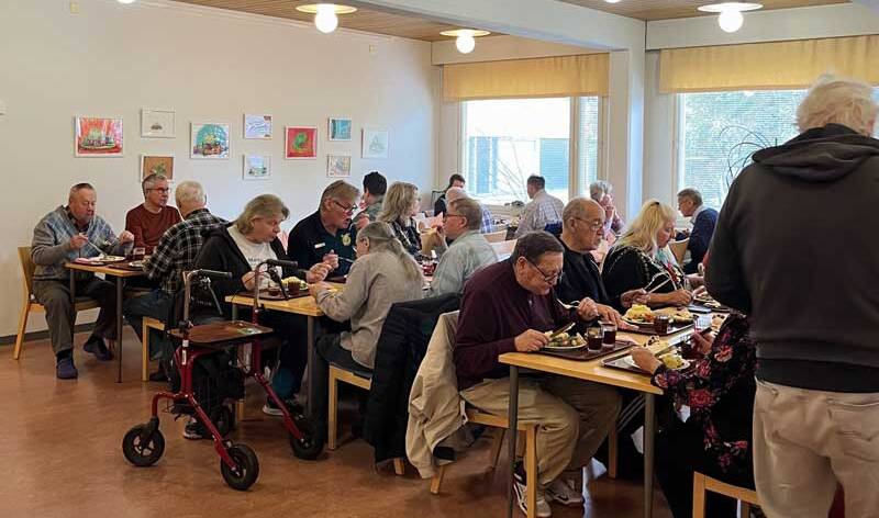 Parikymmentä ruokailijaa istuu kolmen pitkän pöydän ääressä valoissa vaaleassa tilassa.