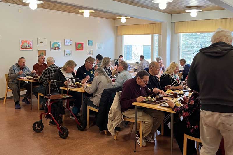Parikymmentä ruokailijaa istuu kolmen pitkän pöydän ääressä valoissa vaaleassa tilassa.