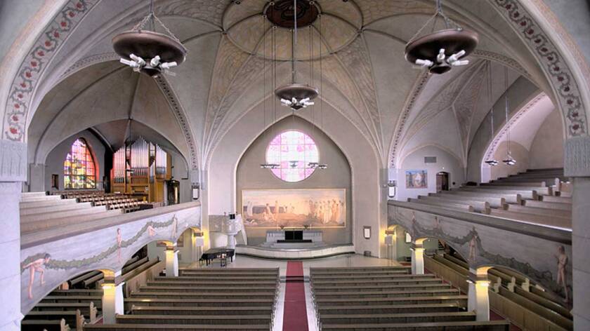 Yleisnäkymä sisältä: Lehteriltä aukeaa näkymä taideteoksin kehystettyyn kirkkosaliin. Kattoholveja koristavat enkelin siivet ja ruusuköynnökset. Lyijylasi-ikkunoista kuultaa pehmeää valoa kirkon sisälle.