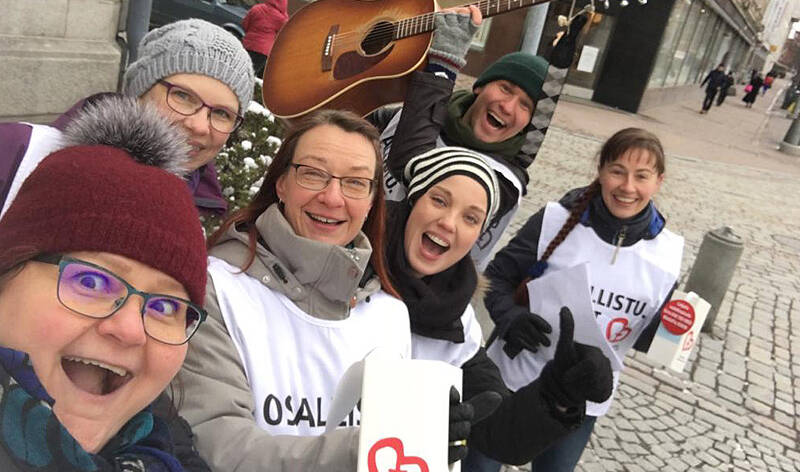 Kuusi henkilö yhteisvastuuliivit päällään ja lippaat kädessään kokoontuneet yhteen iloisen oloisesti.