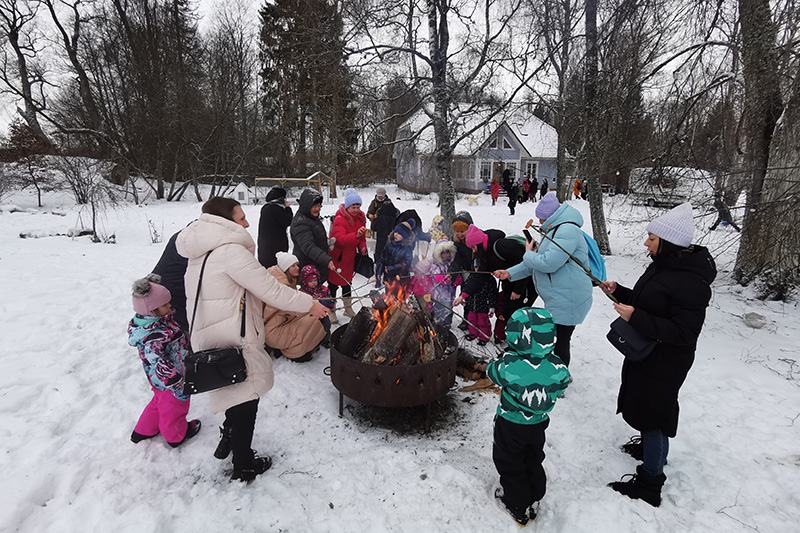 Ulkona lumisessa maisemassa on pata ja siinä nuotio. Lapset ja aikuiset pitävät käsissään keppejä, jotka osoittavat nuotioon.