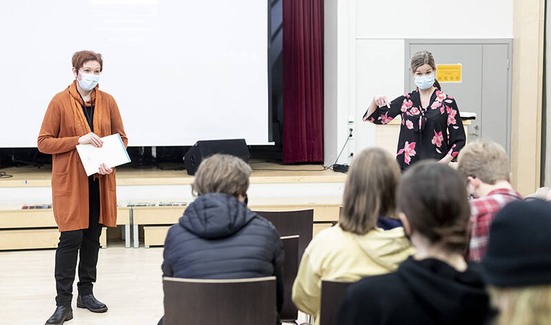 Lukiolaisia nuoria istuu luentosalissa selkä kameraan päin, kun kaksi naista maskit naamalla pitävät esitystä.