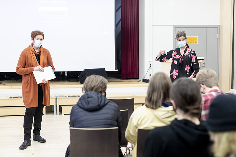 Lukiolaisia nuoria istuu luentosalissa selkä kameraan päin, kun kaksi naista maskit naamalla pitävät esitystä.
