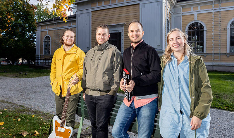 Kolme miestä ja yksi nainen seisoo keltaisen, puisen rakennuksen edustalla nojaten puiston penkkiin. He hymyilevät kameralle, yksi miehistä pitää kädessään kitaraa ja yksi pientä kosketinsoitinta.