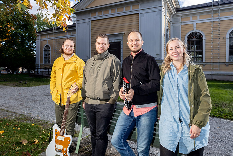 Kolme miestä ja yksi nainen seisoo keltaisen, puisen rakennuksen edustalla nojaten puiston penkkiin. He hymyilevät kameralle, yksi miehistä pitää kädessään kitaraa ja yksi pientä kosketinsoitinta.