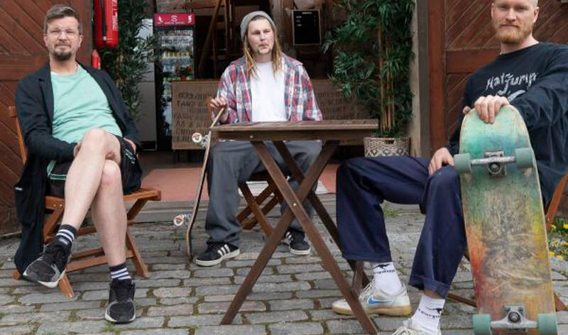 Kolme rennosti pukeutunutta miestä istuu tapulin edustalla terssipöydän ympärillä ja heistä kahdella on skeittiölaudat käsissään.