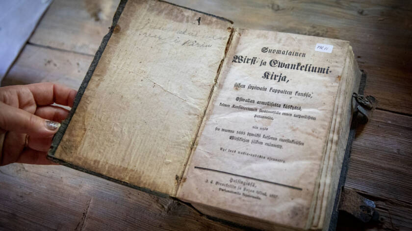 Vanha ja hauras virsikirja on aseteltu kankaan päälle ja käsi pitää sita avoimena kansilehden kohdalta, missä' lukee Suomalainen Wirsi- ja Ewakeliumikirja.