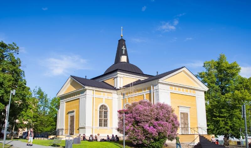 Tampereen keskustorin Vanha kirkko pilvettömänä kesäpäivänä. Ihmisiä istuskelee kirkon edustalla nurmella kiireettömästi.