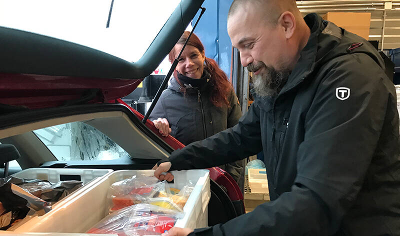Mies on nostanut ison korin täynnä ruokaa autonperäkonttiin ja hymyilee. Nainen seisoo auton vieressä ja hymyilee.