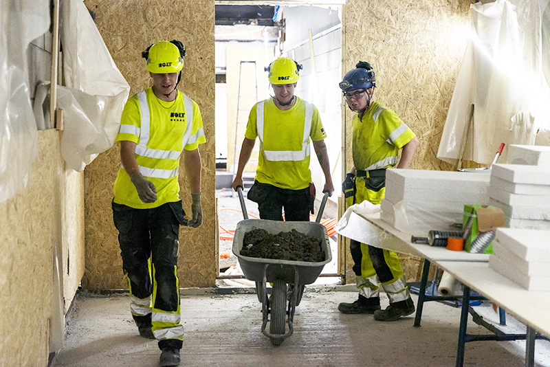 Rakennustyömaalla kulkee kolme henkilöä työvaatteissa. Heistä keskimmäinen työntää kottikärryä.