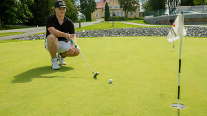 Lippalakkipäinen nuori mies on kyykyssä sileällä kentällä ja pitää käsissään golfmailaa. Hänen edessään on pieni pallo, ja kuvan reunassa lippu.