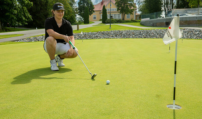 Lippalakkipäinen nuori mies on kyykyssä sileällä kentällä ja pitää käsissään golfmailaa. Hänen edessään on pieni pallo, ja kuvan reunassa lippu.